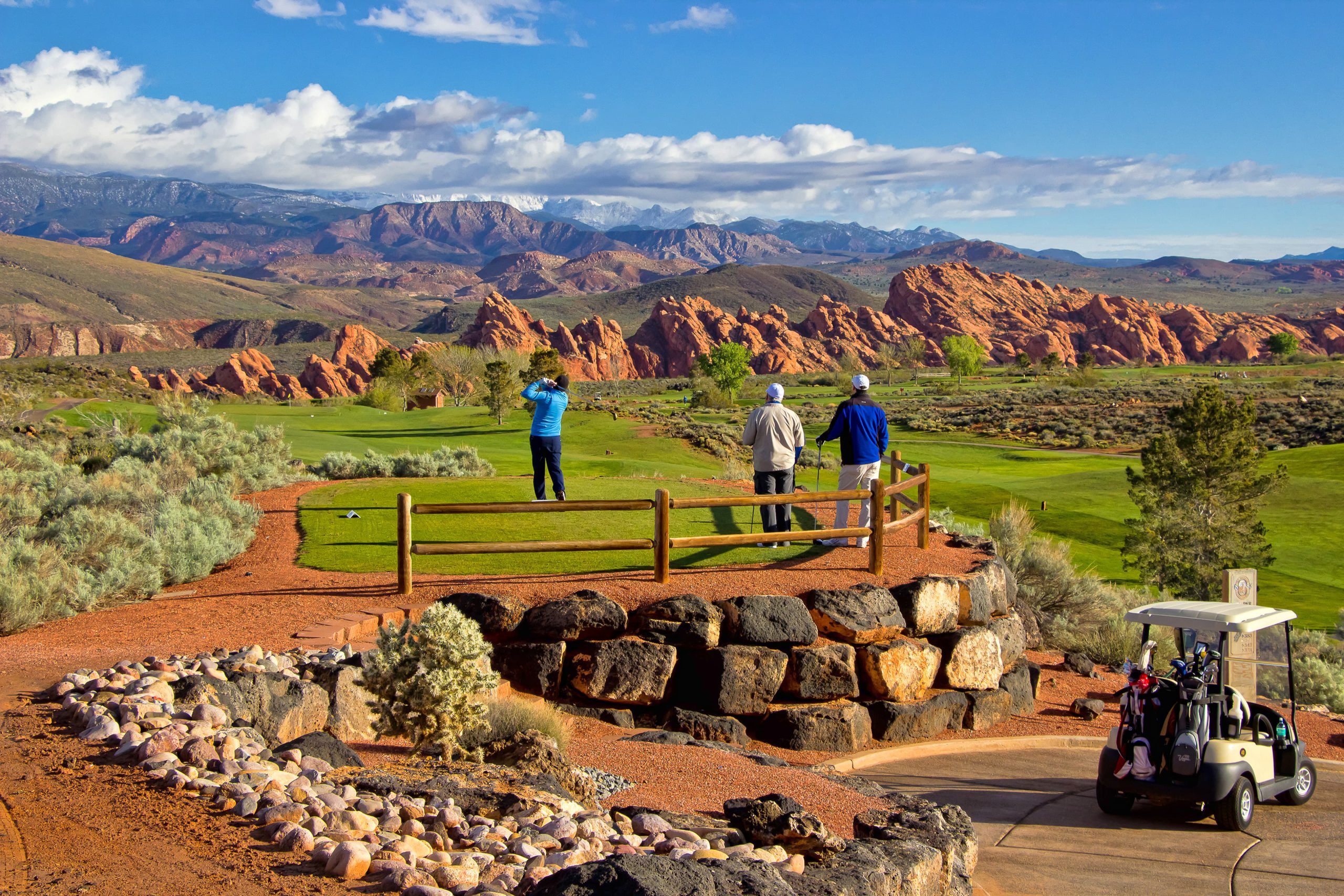 View of Sky Mountain Golf Course. One man takes his swing on the scenic golf course with redrock in the background as two men look on from the foot path.