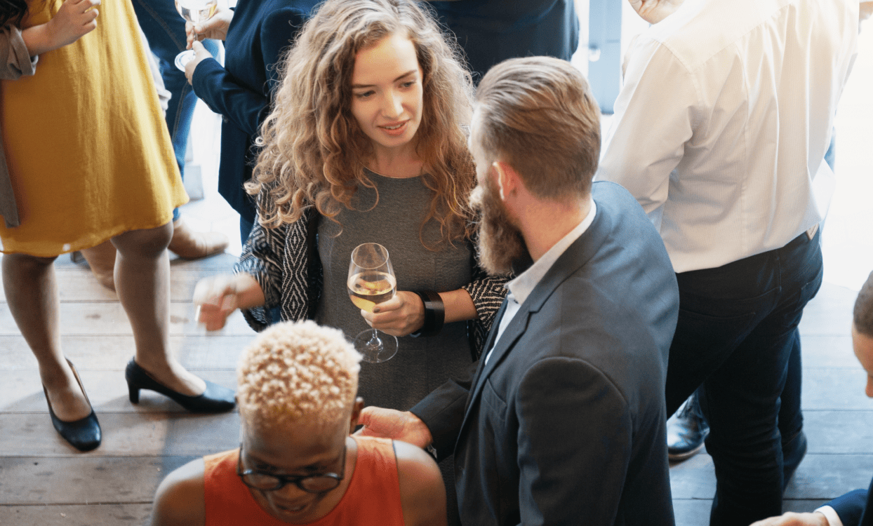 Overview of group of people socializing at a party. A woman with curly hair holds a wine glass with white wine while talking to a man in a suit and tie
