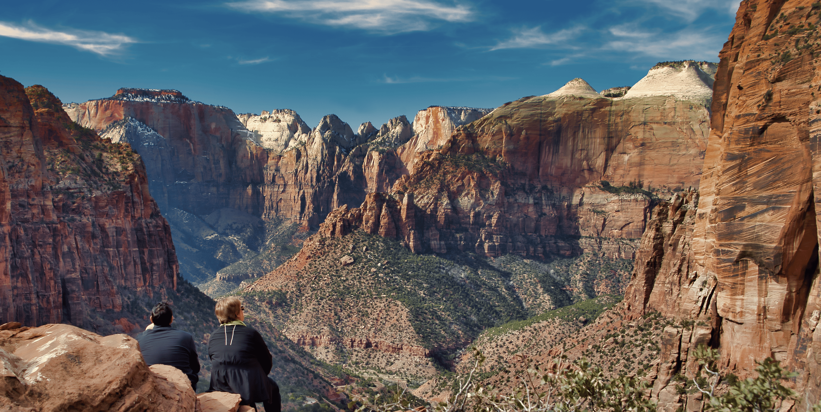 Two people sit looking out towards a magical red rock canyon that sprawls for miles into the distance.