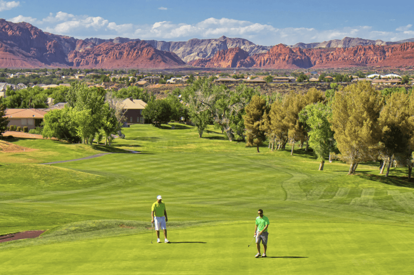 Two golfers in green shirts and tan pants stand on a manicured green just after the fairway. Picturesque red rock mountains loom in the distance