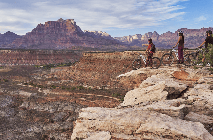 Three mountain bikers stand on an overhang overlooking a red rock canyon. A road trails off in the distance, disappearing behind a rock formation